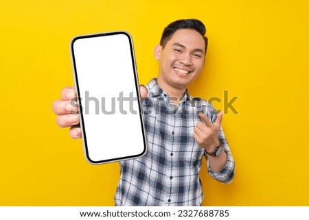 Smiling handsome young Asian man wearing a white checkered shirt pointing finger at mobile phone with blank screen, Advertising new mobile app isolated over yellow background. People Lifestyle Concept