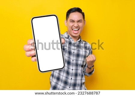 Smiling handsome young Asian man wearing a white checkered shirt showing a screen mobile phone for an advertisement and celebrating success isolated over yellow background. People Lifestyle Concept