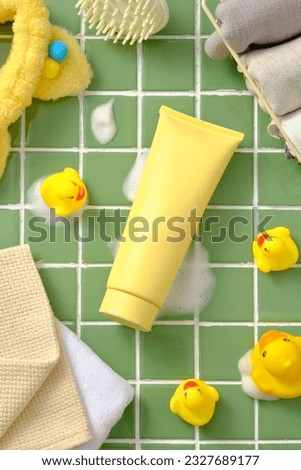 Unbranded plastic tube decorated with toy yellow duck, towel and hair tier, mirror and scalp massage brush on green ceramic background. Mockup of skin care cosmetic tube of beauty facial