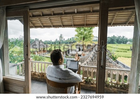 A successful middle-aged man of European appearance works on a laptop while sitting in a villa with a gorgeous view of the rice fields.