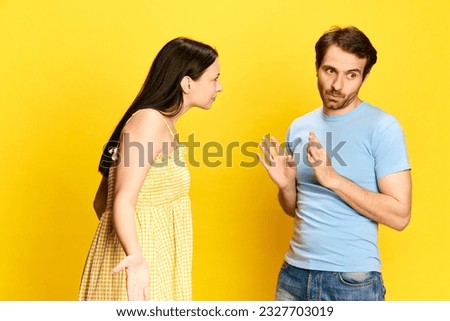 Young woman emotionally talkin to man, asking questions against yellow studio background. Man making excuses. Concept of friendship, relationship, communication, emotions, lifestyle, ad Royalty-Free Stock Photo #2327703019