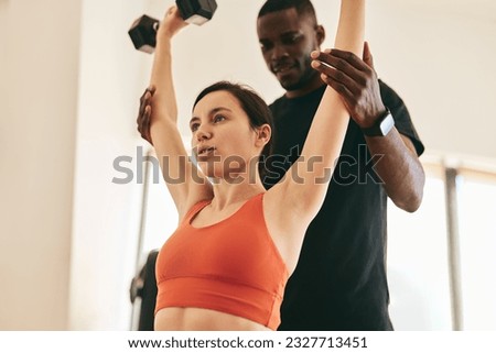 Crop black personal instructor helping young fit female doing exercise with dumbbells and arms raised during fitness training at gym