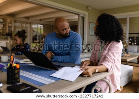 Biracial parents using laptop, looking at bills and talking at table with daughter in background. Finance, family, communication, lifestyle and domestic life, unaltered.