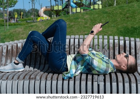 Woman lying down on park bench and flip through display on mobile phone. Young adult use smart phone for chatting. Fingers touch the screen of telephone. Female resting and communication on cellphone.