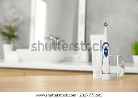 Electric toothbrush, tube with paste and glass of water on wooden table in bathroom. Space for text