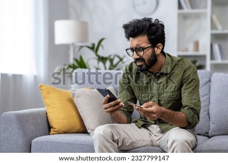 Upset man at home cheated and denied money transfer, hispanic sitting on sofa at home with phone and bank credit card, cheated and disappointed in living room. Royalty-Free Stock Photo #2327799147