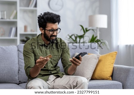 Upset man at home cheated and denied money transfer, hispanic sitting on sofa at home with phone and bank credit card, cheated and disappointed in living room. Royalty-Free Stock Photo #2327799151