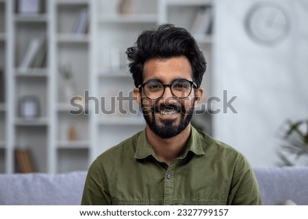 Close-up portrait of young smiling hispanic man smiling and looking at camera while sitting on sofa in living room.