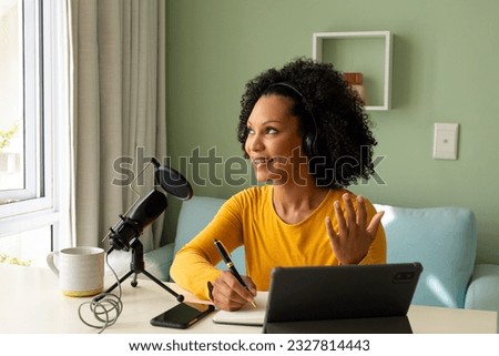 Happy biracial woman using headphones, tablet and microphone podcasting from home. Communication, technology, social media and domestic life, unaltered.