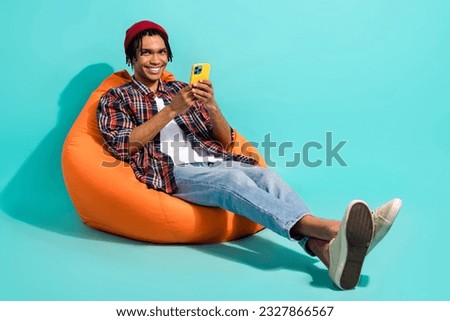 Full size photo of positive guy dressed checkered shirt jeans sit on pouf read post on smartphone isolated on teal color background