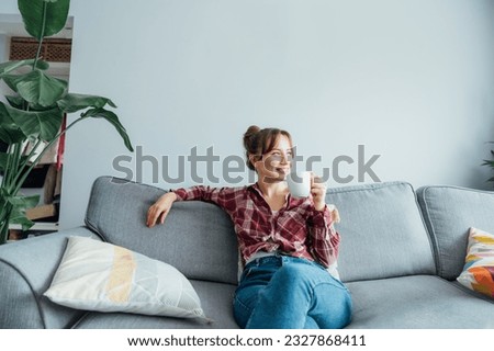 Young smiling woman sitting on sofa and looking away while drinking coffee or tea. Young brunette woman relaxing after housekeeping, home cleaning. Portrait of relaxed female resting at home. Royalty-Free Stock Photo #2327868411