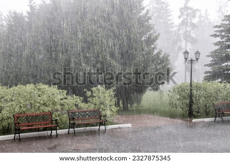 Rain with hail on a summer day. Large puddles on the sidewalk during heavy rain. Heavy rain in the park. Wet benches on a paved path. Spray drops and bubbles in large puddles. Royalty-Free Stock Photo #2327875345