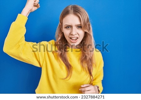 Young caucasian woman standing over blue background angry and mad raising fist frustrated and furious while shouting with anger. rage and aggressive concept.  Royalty-Free Stock Photo #2327884271