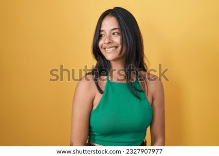 Brunette woman standing over yellow background looking away to side with smile on face, natural expression. laughing confident. 