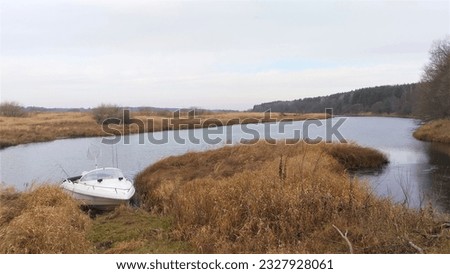 Fishing on the river in late autumn. A plastic motorboat is moored to a grassy bank among dry reeds In the boat is a spinning rod and a net for lifting fish. A forest stands on the high shore Overcast Royalty-Free Stock Photo #2327928061