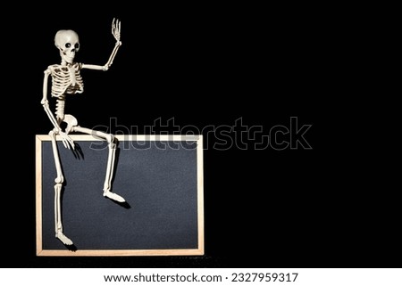 Funny human skeleton sitting on an empty black flipchart on a black background, copy space, mockup.