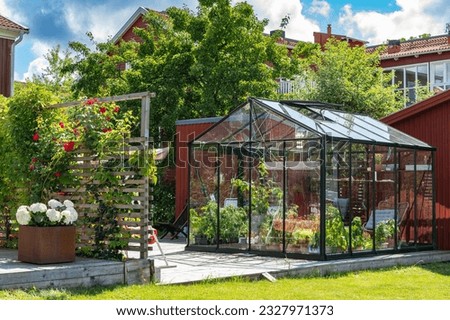 Beautiful greenhouse glass house in the garden yard near the villa. Wicker rattan chairs inside. Lots of pots with different plants.  Greenhouse for growing plant seedlings. Landscape garden design. Royalty-Free Stock Photo #2327971373