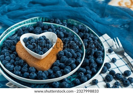 Fresh bilberries on top in heart shaped plate on bilberries background. A lot of fresh bilberries in a blue bowl on bilberries background. High quality photo