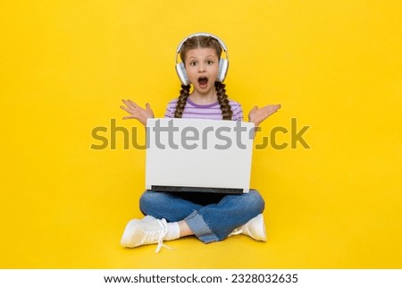 A child with a laptop. A young girl in headphones opened her mouth in shock from surprise and spread her hands in different directions. Internet communication for teenagers. Yellow isolated background