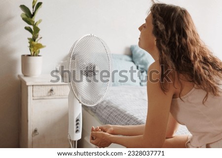 Caucasian woman sits in front of an air conditioner fan and cools down in summer heat, during strong heat wave at home, heat stroke, malaise from temperature in summer, indoor ventilation Royalty-Free Stock Photo #2328037771