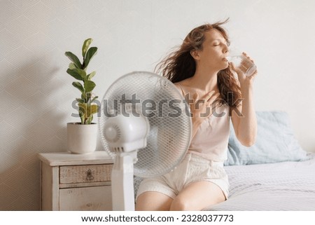 young European woman cools down at home with help of an air conditioner fan and drinks cool water with ice sitting on bed in front of an electric ventilator during intense heat of house, heatstroke Royalty-Free Stock Photo #2328037773
