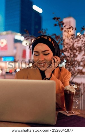 In the enchanting ambiance of a nocturnal cityscape, a hijab-clad girl engrossed in her laptop creates a mesmerizing image, embodying the fusion of technology, empowerment, and the vibrant urban