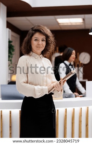 Administrator carrying record files and standing near front desk in hotel lobby, verifying service as manager. Female supervisor checking booking register and welcoming people. Royalty-Free Stock Photo #2328045719