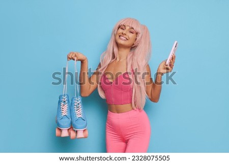 Pretty woman wearing Barbie style, posing on blue background in pink clothes with pink hair, holding roller-skates and phone in hands and smiles, Barbie fashion concept, copy space Royalty-Free Stock Photo #2328075505