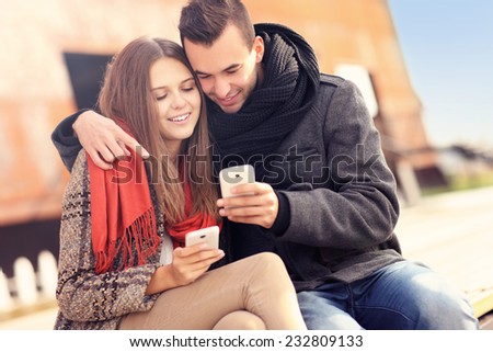 A picture of a young couple sitting on a bench and using smartphones on an autumn day