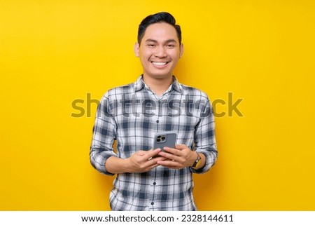 Smiling satisfied handsome young Asian man wearing a white checkered shirt holding cellphone isolated over yellow background. People Lifestyle Concept