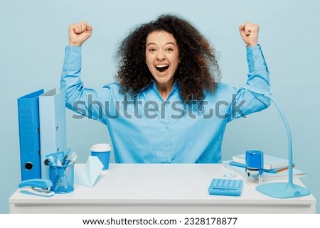 Young fun employee business woman wear casual shirt sit work at white office desk doing winner gesture celebrate clenching fists say yes isolated on plain pastel light blue background studio portrait