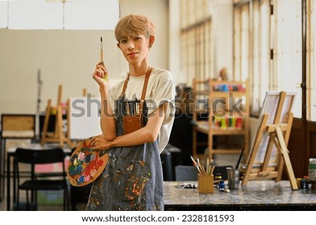Portrait of asian gay man student wearing apron holding paintbrush and looking at camera