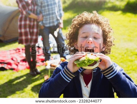 Enthusiastic boy taking large bite of hamburger at sunny campsite