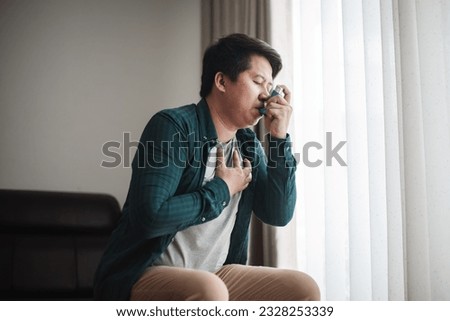 Panoramic shot of asthmatic man using inhaler with spacer. Fat man using asthma inhaler while sitting at home due to allergy. Asia man using spray asthma. Royalty-Free Stock Photo #2328253339