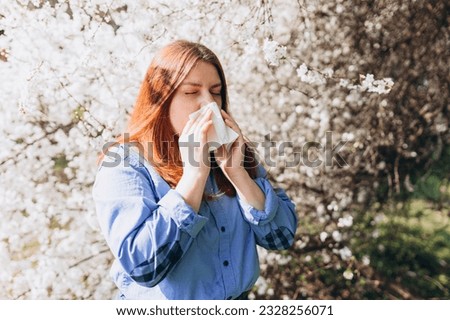 Sneezing young redhead woman with nose wiper among blooming trees in park. Portrait of sick women sneezes in white tissue, suffers from rhinitis and running nose. Symptoms of cold or allergy. Royalty-Free Stock Photo #2328256071