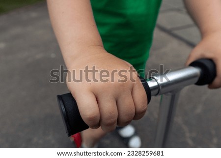 Child's hands are holding the steering wheel of a scooter. The boy is riding a scooter