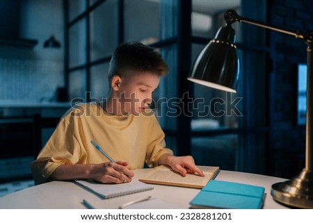 Focused redhead pupil student boy studying at home writing in exercise book doing homework, learning sitting at table under light of lamp at night. Clever schoolkid reading textbook sitting at desk. Royalty-Free Stock Photo #2328281201