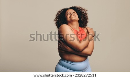 Body confident woman embracing her body, expressing self-love and self-acceptance. Young plus-size woman standing in a studio in fitness clothing, embracing her natural physique with joy. Royalty-Free Stock Photo #2328292511