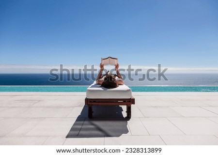 Woman reading on lounge chair at poolside overlooking ocean Royalty-Free Stock Photo #2328313899