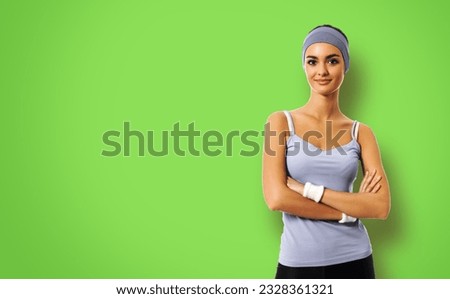 Portrait of smiling sporty brunette woman in grey sportswear, isolated against green background. Young female fitness instructor or personal trainer in crossed arms pose at studio.