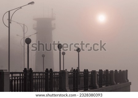 An old lighthouse in the fog on a renovated embankment with lanterns. The disk of the sun with a red halo. Blagoveshchensk, Far East, Russia.