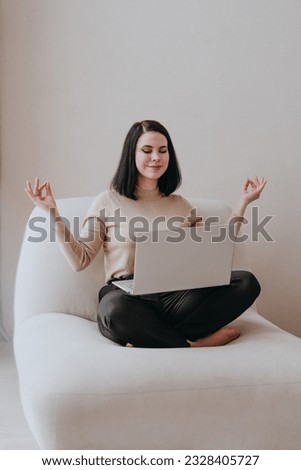 Young smiling brunette woman with closed eyes in business clothes siting barefoot in a lotus position with a laptop indoor. Balance of work and mental health concept. Royalty-Free Stock Photo #2328405727