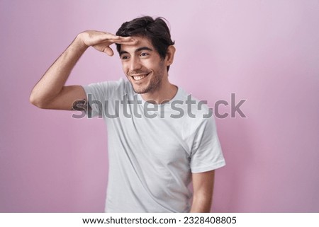 Young hispanic man standing over pink background very happy and smiling looking far away with hand over head. searching concept.  Royalty-Free Stock Photo #2328408805