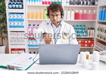 Hispanic young man working at pharmacy drugstore working with laptop pointing with finger to the camera and to you, confident gesture looking serious 