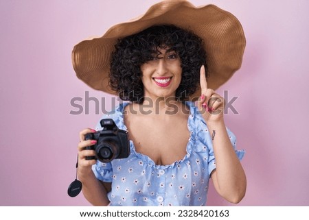 Young brunette woman with curly hair using reflex camera surprised with an idea or question pointing finger with happy face, number one 