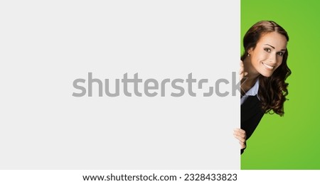 Happy excited woman in black confident suit, peek out showing blank white banner signboard with copy space for sign text. Business and ad concept. Green color background.