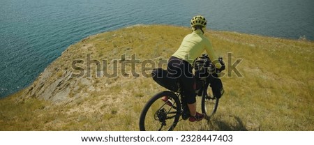 The beautiful woman is ride on top of a hill with the sea in the background, near the canyon. The sporty girl travels on a bicycle with bikepacking bags. The traveler journey on touring bicycle