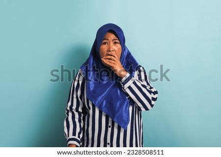 A shocked middle-aged Asian woman in a blue hijab is watching television, pointing the TV remote controller towards the camera, and switching channels with an open mouth, Isolated on a blue background