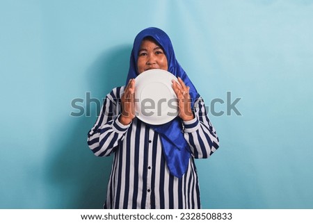 An excited middle-aged Asian woman in a blue hijab and a striped shirt is standing and holding an empty white plate. She is isolated on a blue background