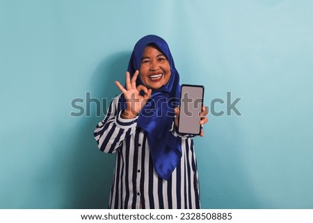 An excited middle-aged Asian woman in a blue hijab and a striped shirt is holding her phone with a blank white screen while making an approval sign or OK gesture. She is isolated on a blue background.